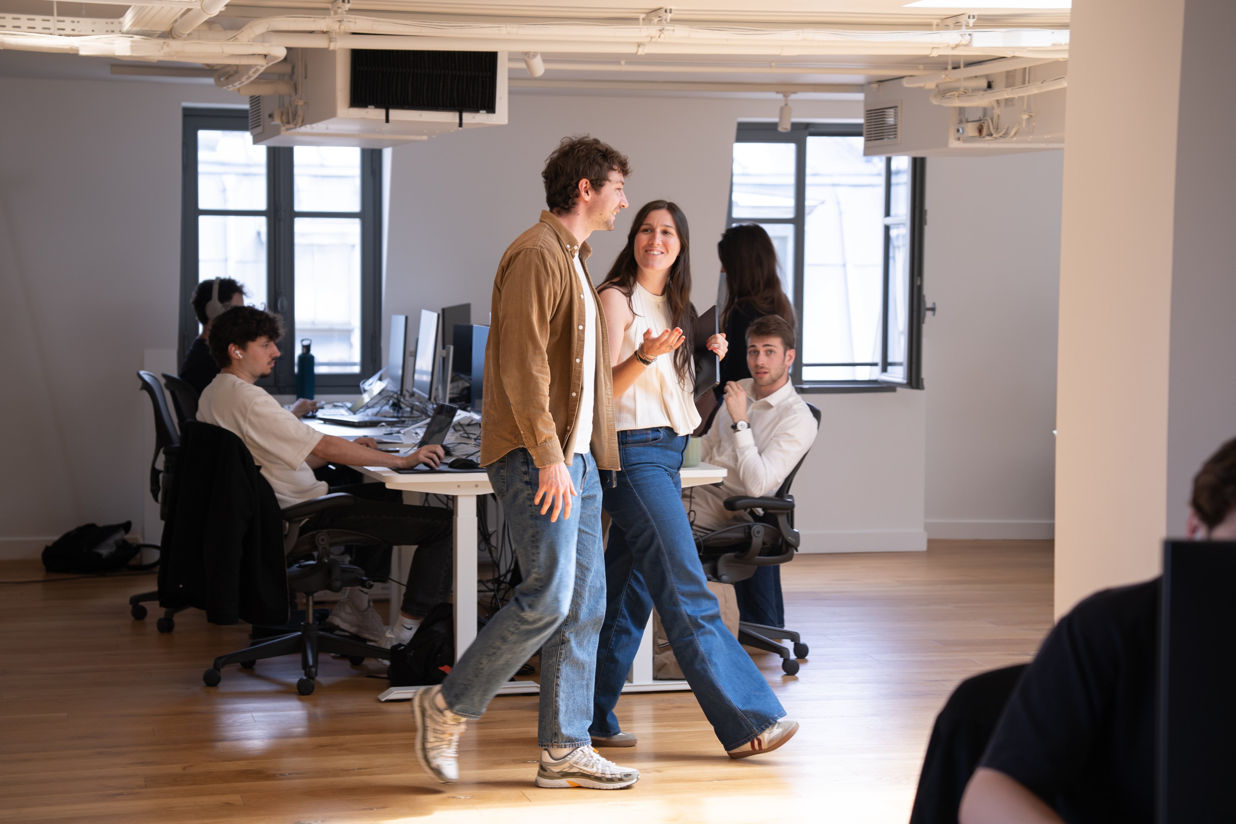 Trois collaborateurs debout discutent dans un open space lumineux, deux autres travaillant à leurs postes en arrière-plan.